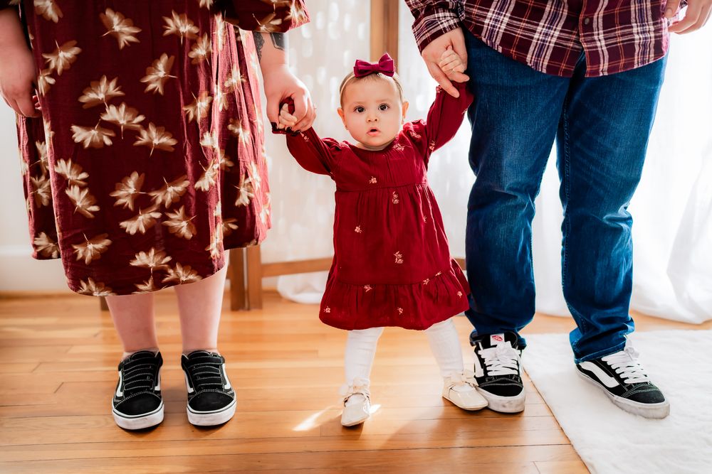 A small child in a red dress stands between two people wearing casual clothing on a hardwood floor.