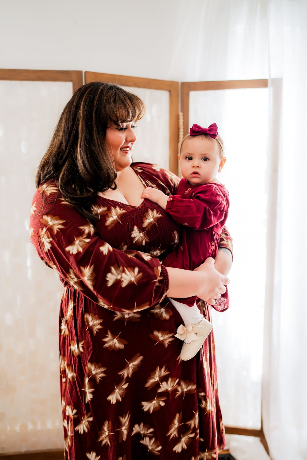 A woman in a burgundy floral dress holds a baby wearing a red outfit and bow against a white background.