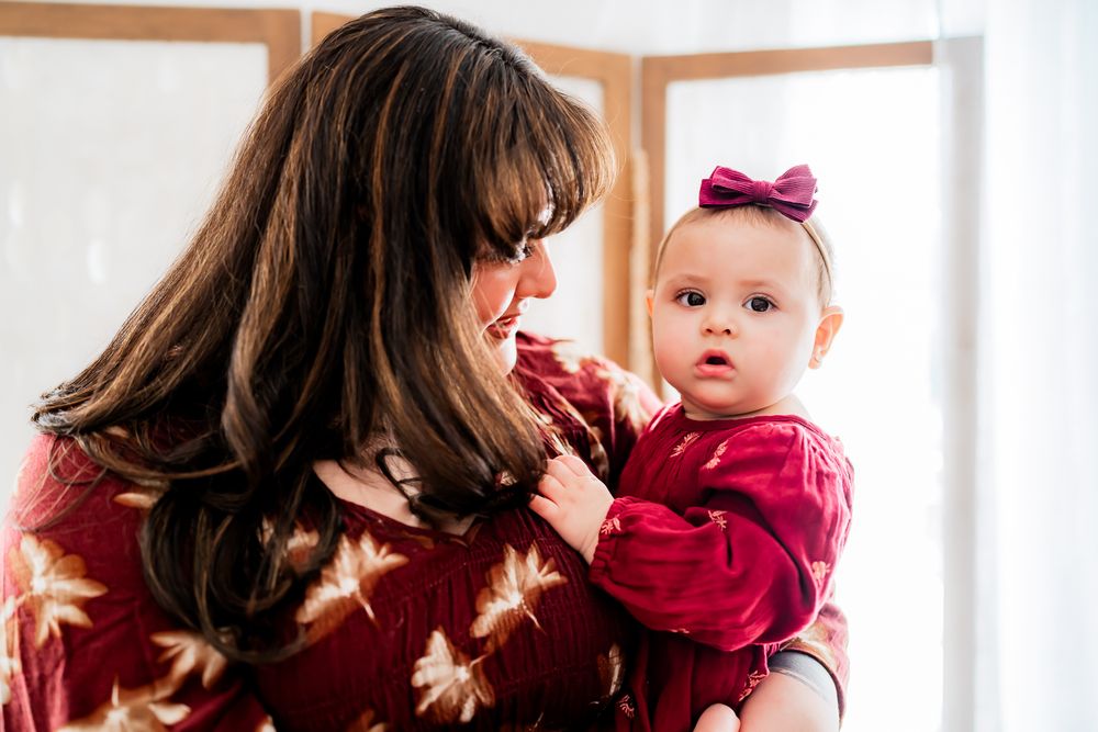 Parent and baby share a tender moment by a bright window while wearing matching burgundy sweaters.