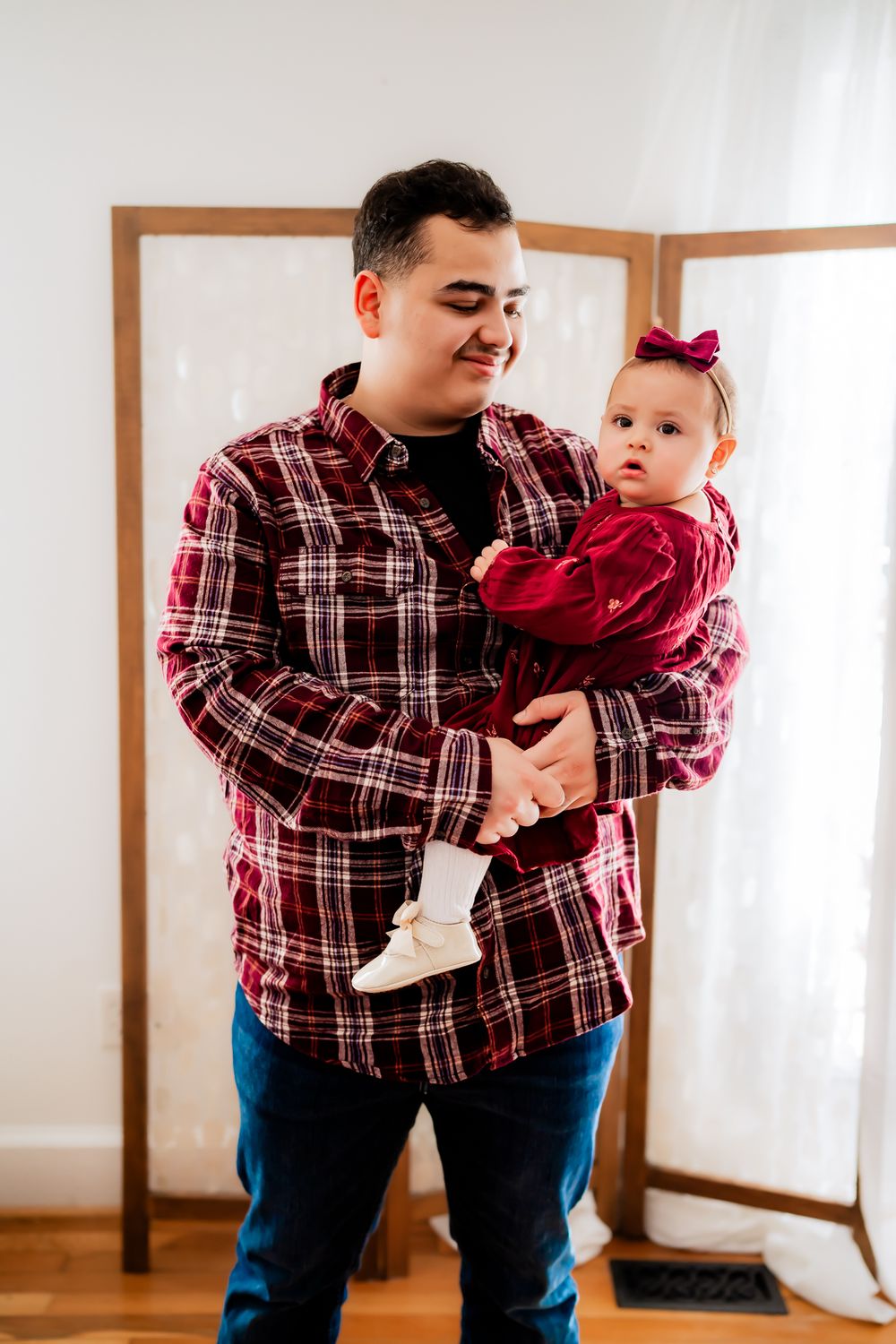 Parent and baby wearing matching red plaid flannel shirts share a tender moment in a bright indoor setting.