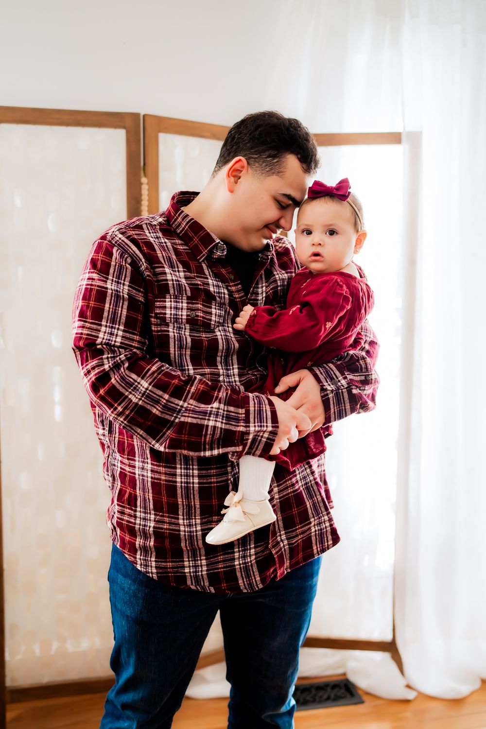 Parent and baby in coordinating red plaid shirts interact playfully in front of white curtains.