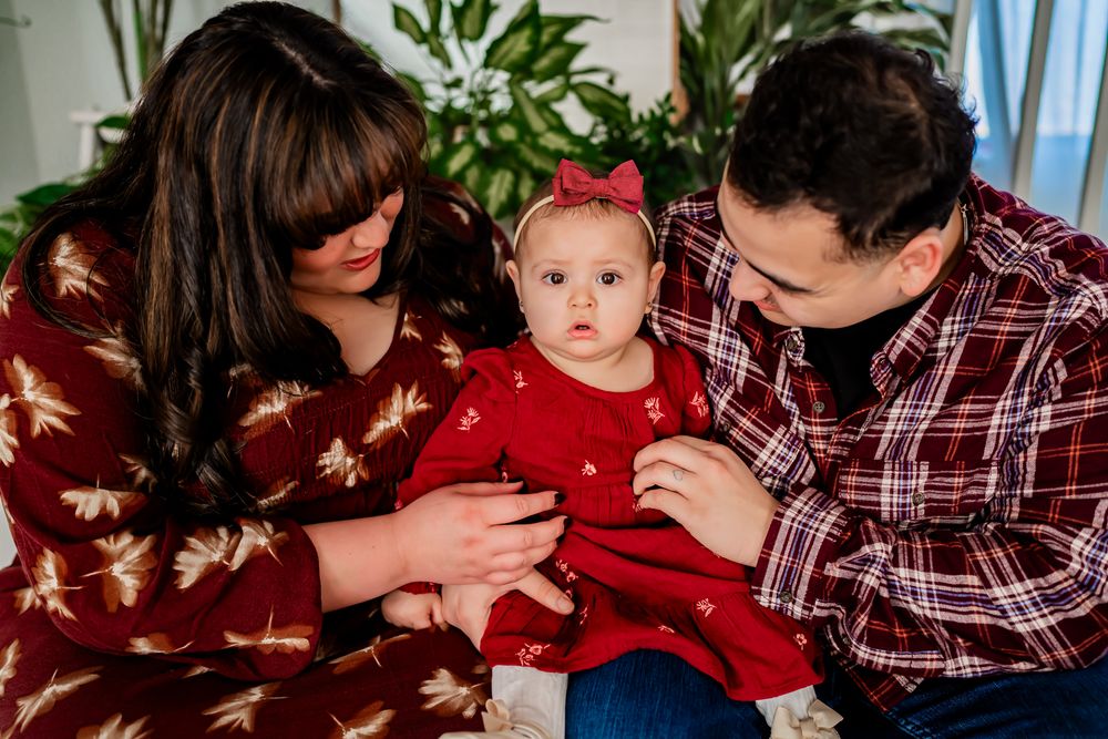 A baby in a red holiday dress is held by two adults wearing coordinating outfits indoors.