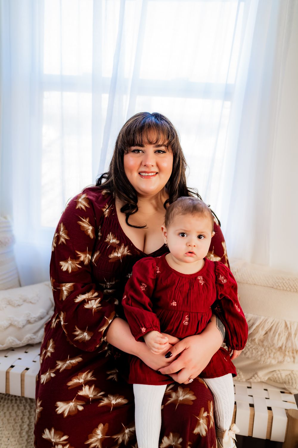 Mother and baby in matching burgundy sweaters pose together on white furniture in a bright sunlit room.