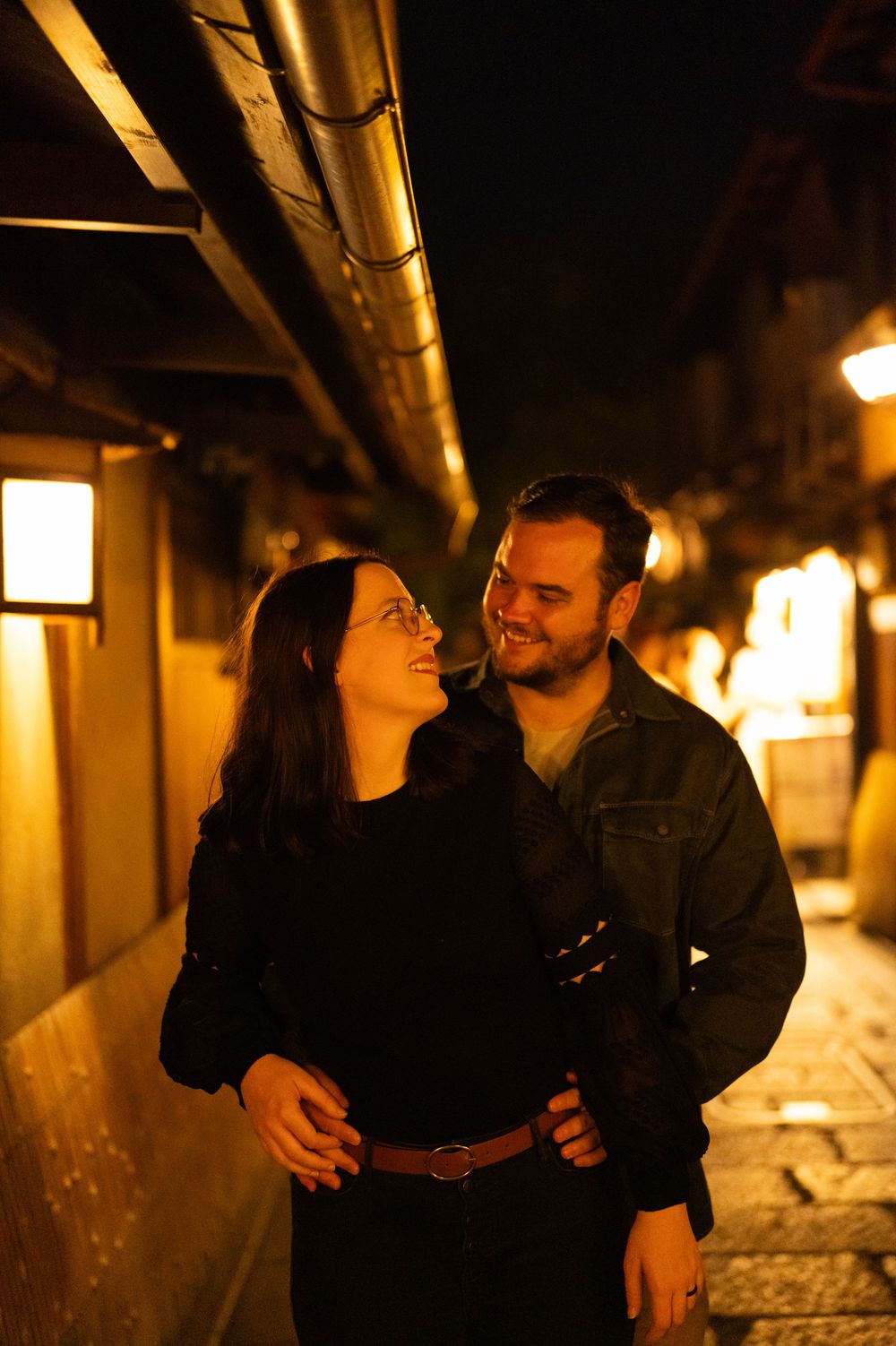 Smiling couple embracing on a warmly lit traditional Japanese street at night in Kyoto.
