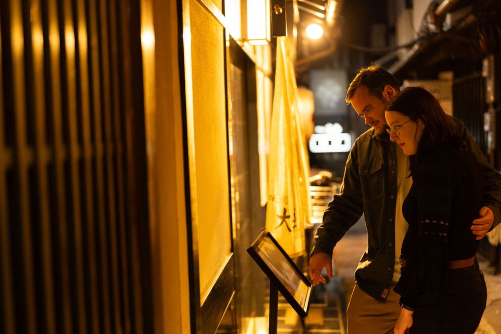 Couple reading a menu outside a glowing yellow Japanese restaurant storefront at night.