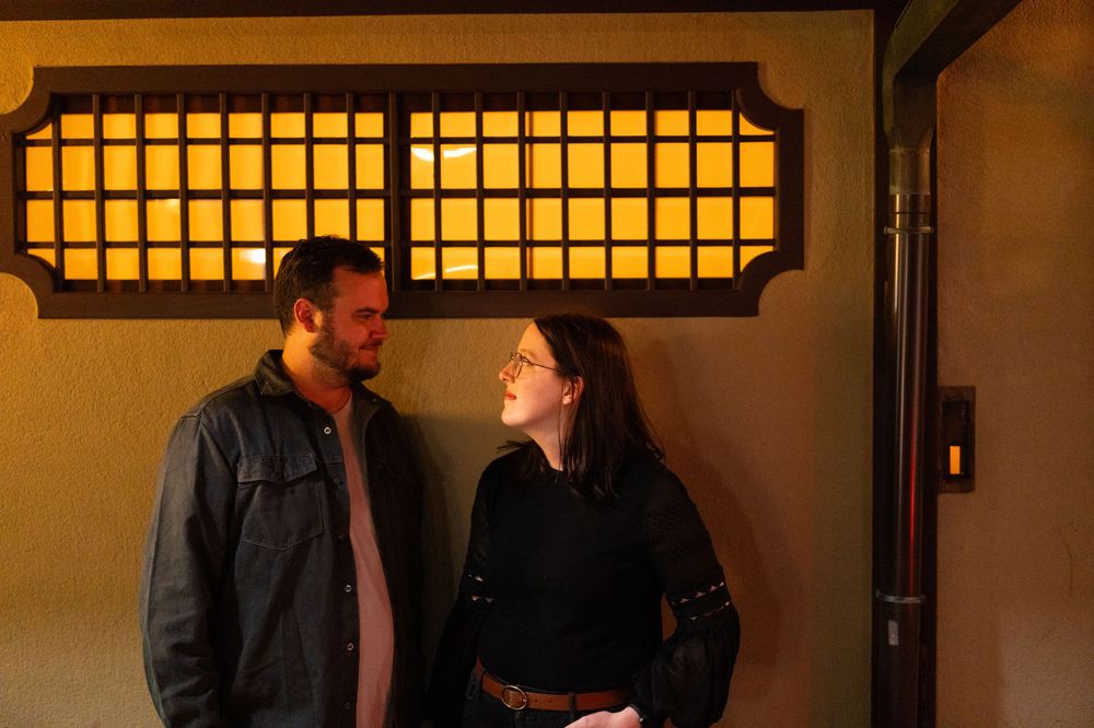 Couple sharing a smile standing before a glowing traditional Japanese lattice window at night.