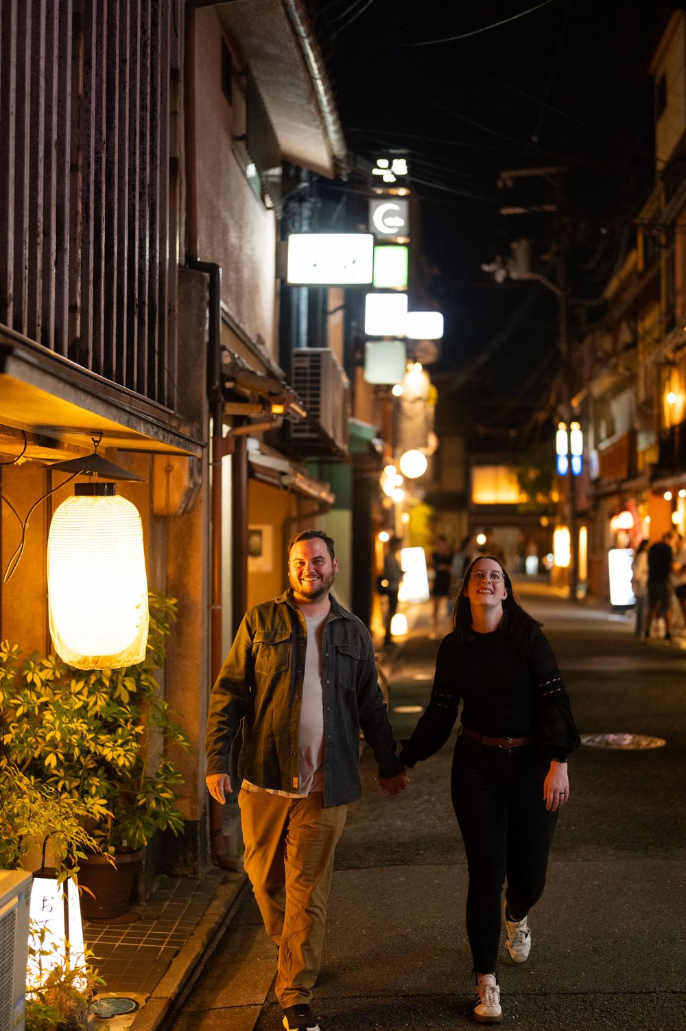 Happy couple holding hands walking down a lantern-lit Japanese street at night in Kyoto.