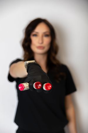A person wearing a black t-shirt poses in a portrait series with red lipstick tubes arranged on their shoulder.