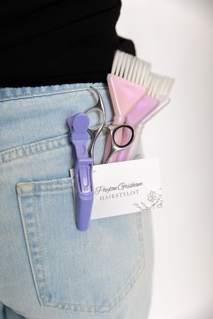 Hairstyling tools including scissors, combs, and a purple clip tucked into a light denim jeans pocket.