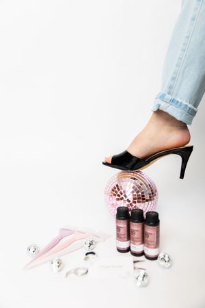 Black high heel mule shoe stepping on pink disco ball with beauty products and decorative elements on white background.