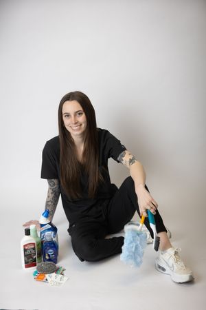 Person in black clothing sits on white background with various cleaning supplies and products arranged around them.