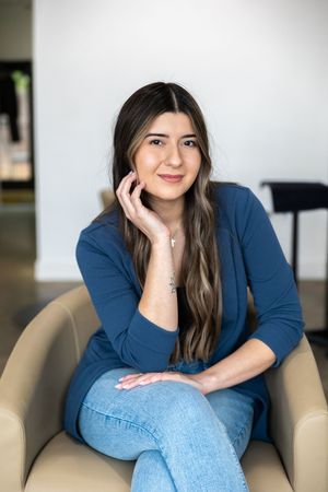 A woman in a navy blue sweater and jeans sits casually on a beige armchair in a minimalist setting.