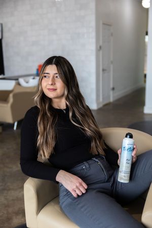 Person in black sweater relaxes on beige chair while holding a white bottle in modern minimalist space.