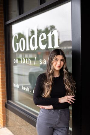 Woman in black top poses against storefront window with 'Golden' signage in modern salon setting.