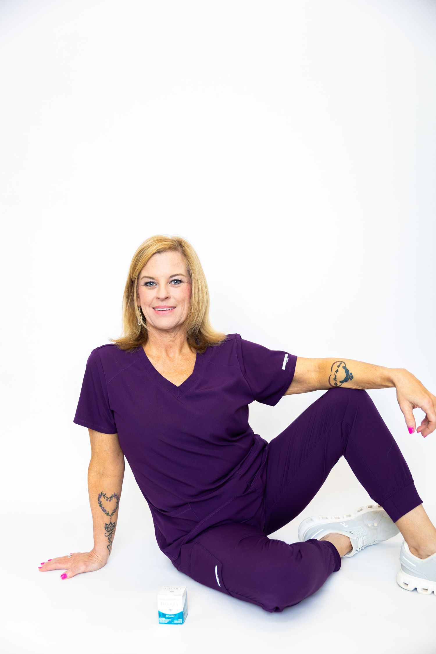 Woman in purple workout attire sitting on floor against white background with visible tattoos and pink nails.