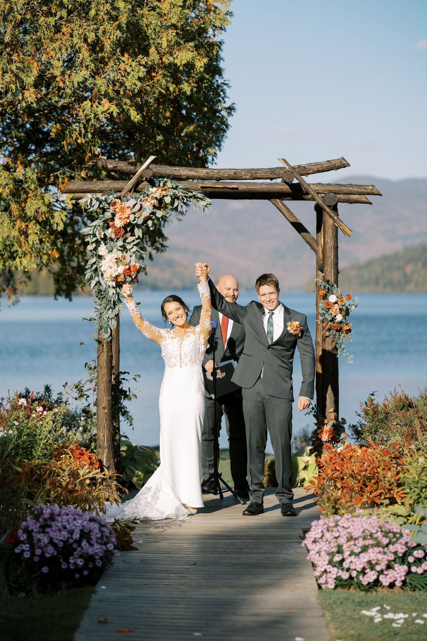 Scenic lakeside wedding ceremony on wooden deck with mountain views and colorful autumn flowers in bloom.