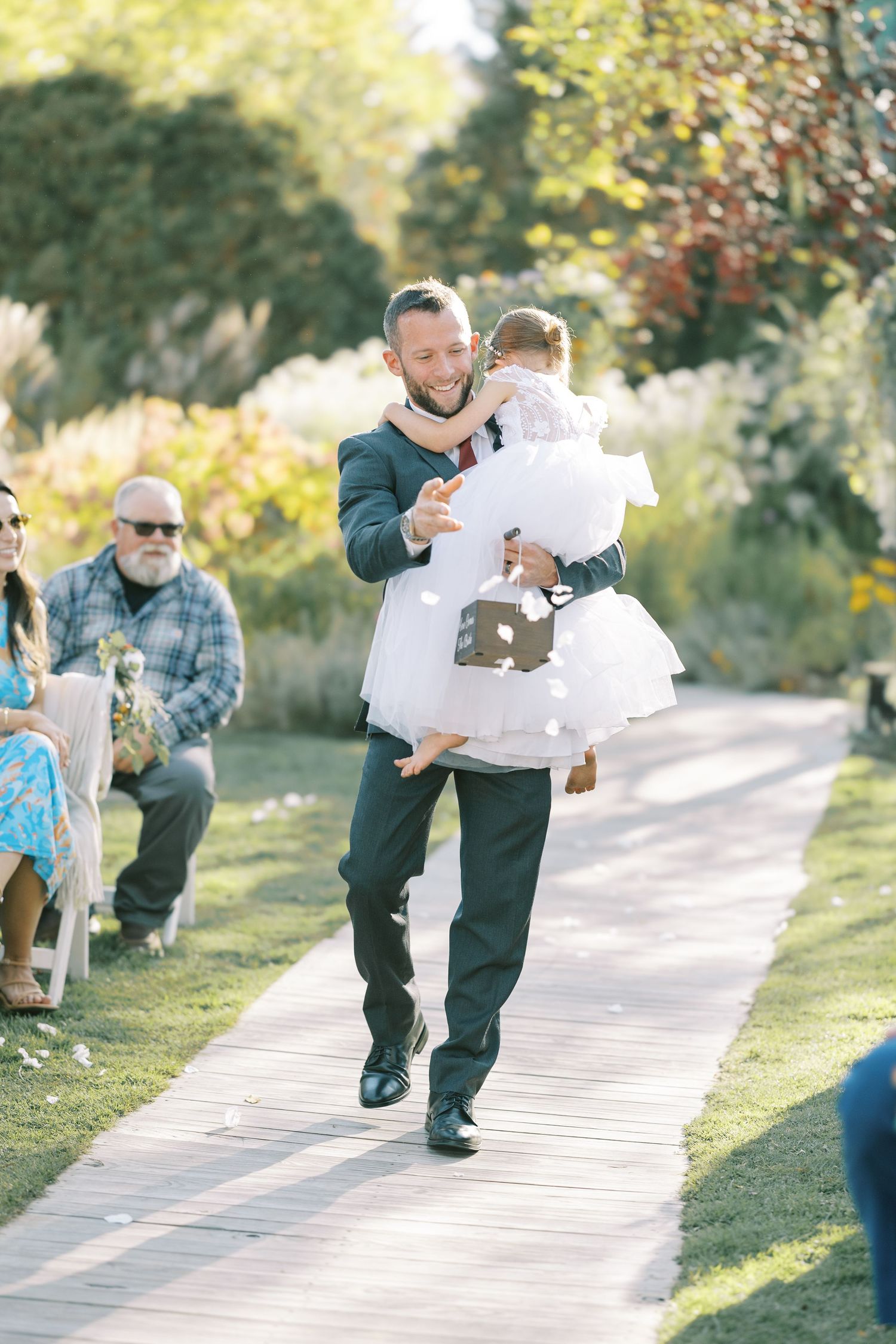 A sequence of photos showing a ring bearer in a suit carrying a white pillow down an outdoor garden path at a wedding.