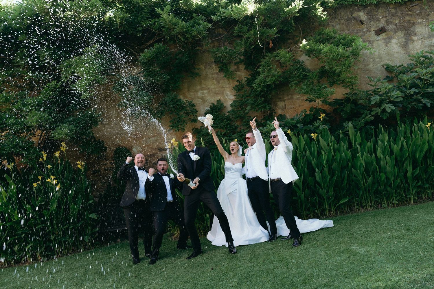 A person in a formal black tuxedo and white shirt poses in a garden setting with white flowers in the background.
