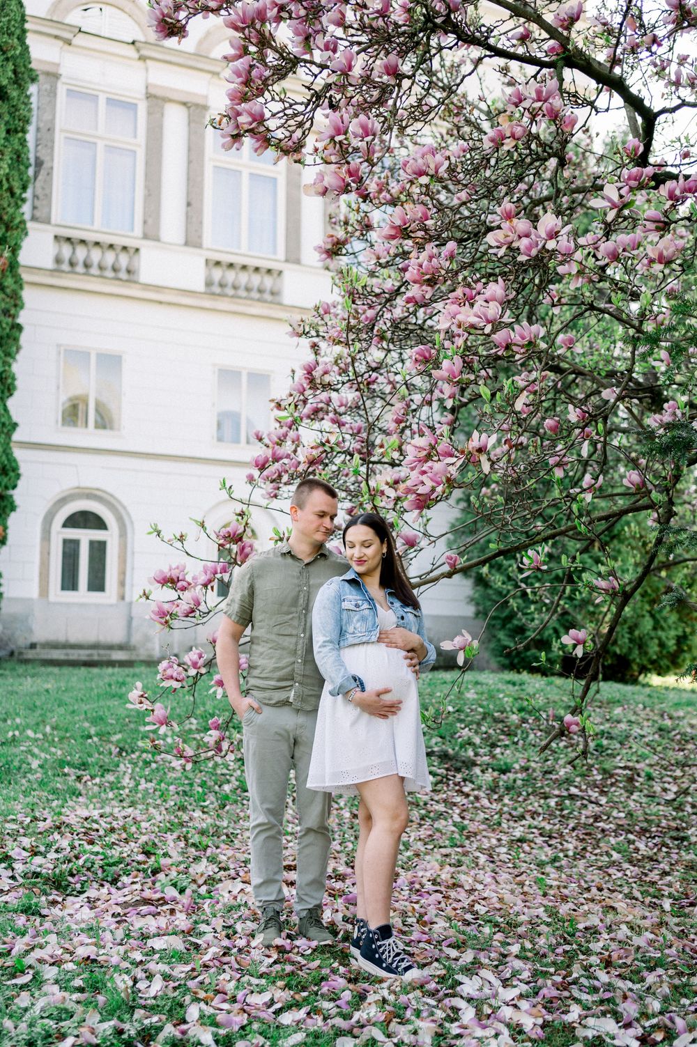 A couple poses together beneath flowering magnolia trees near a white building.