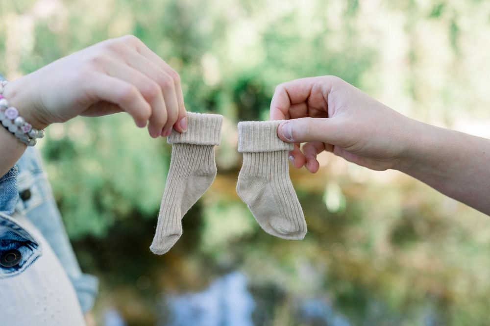 Hands hold tiny white baby socks against a blurred natural background.