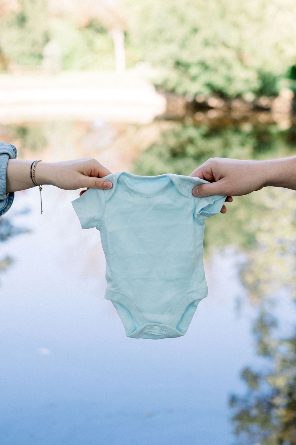 Hands holding a mint green baby onesie against a blue water backdrop.