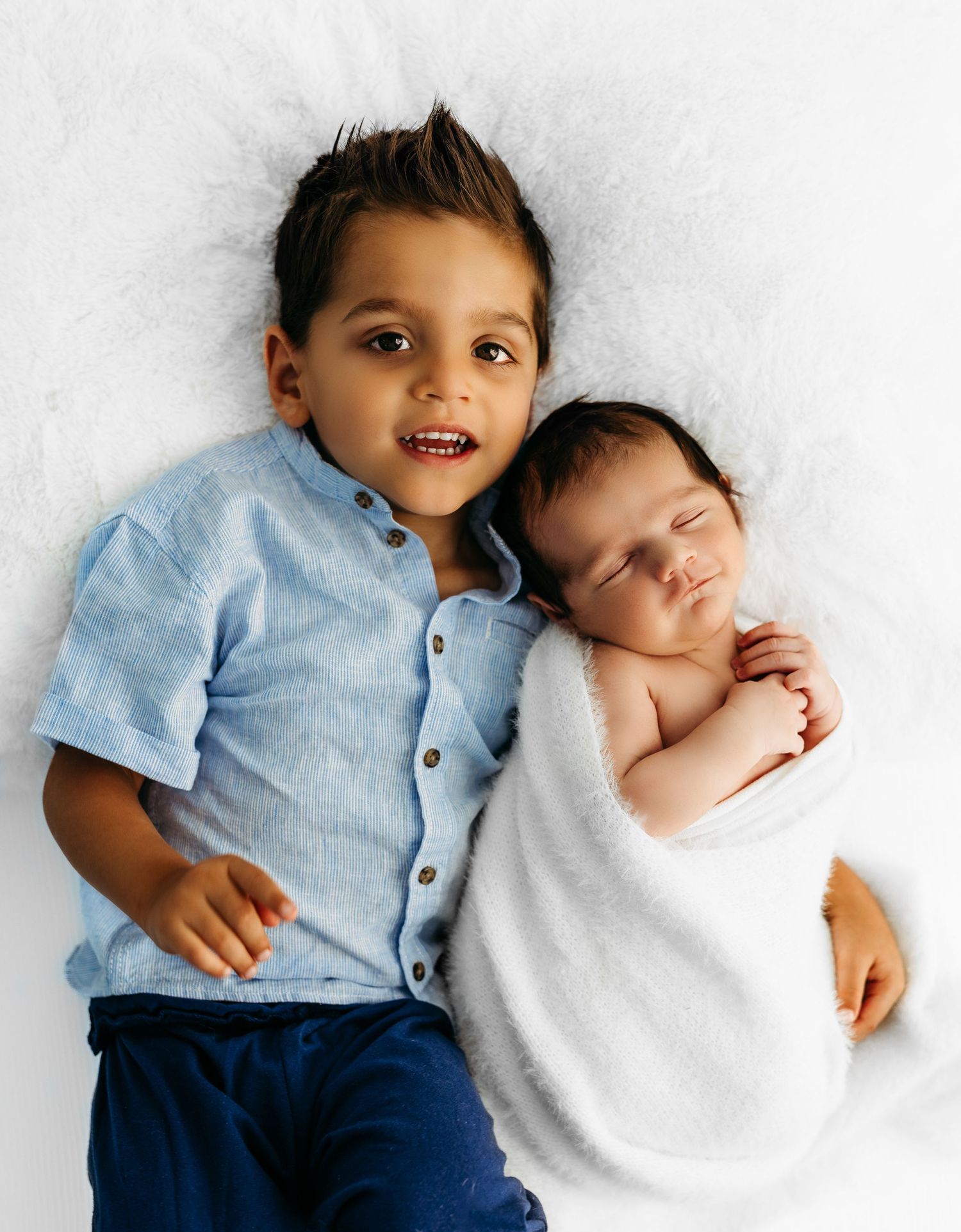 A newborn baby wrapped in white swaddle lies next to a child and black and white dog on fluffy white bedding.