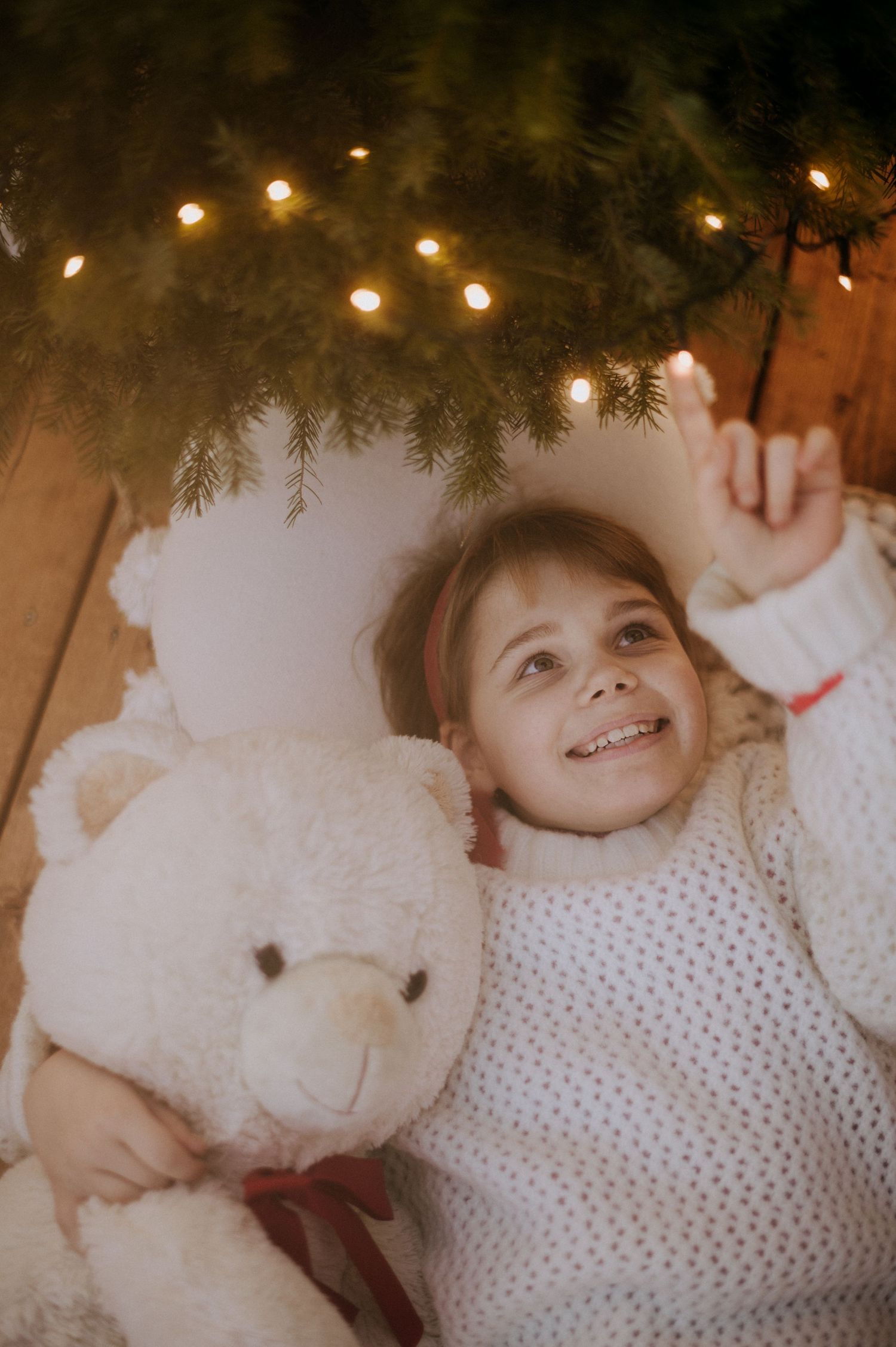 A child lies on white bedding beneath twinkling holiday lights and evergreen branches.