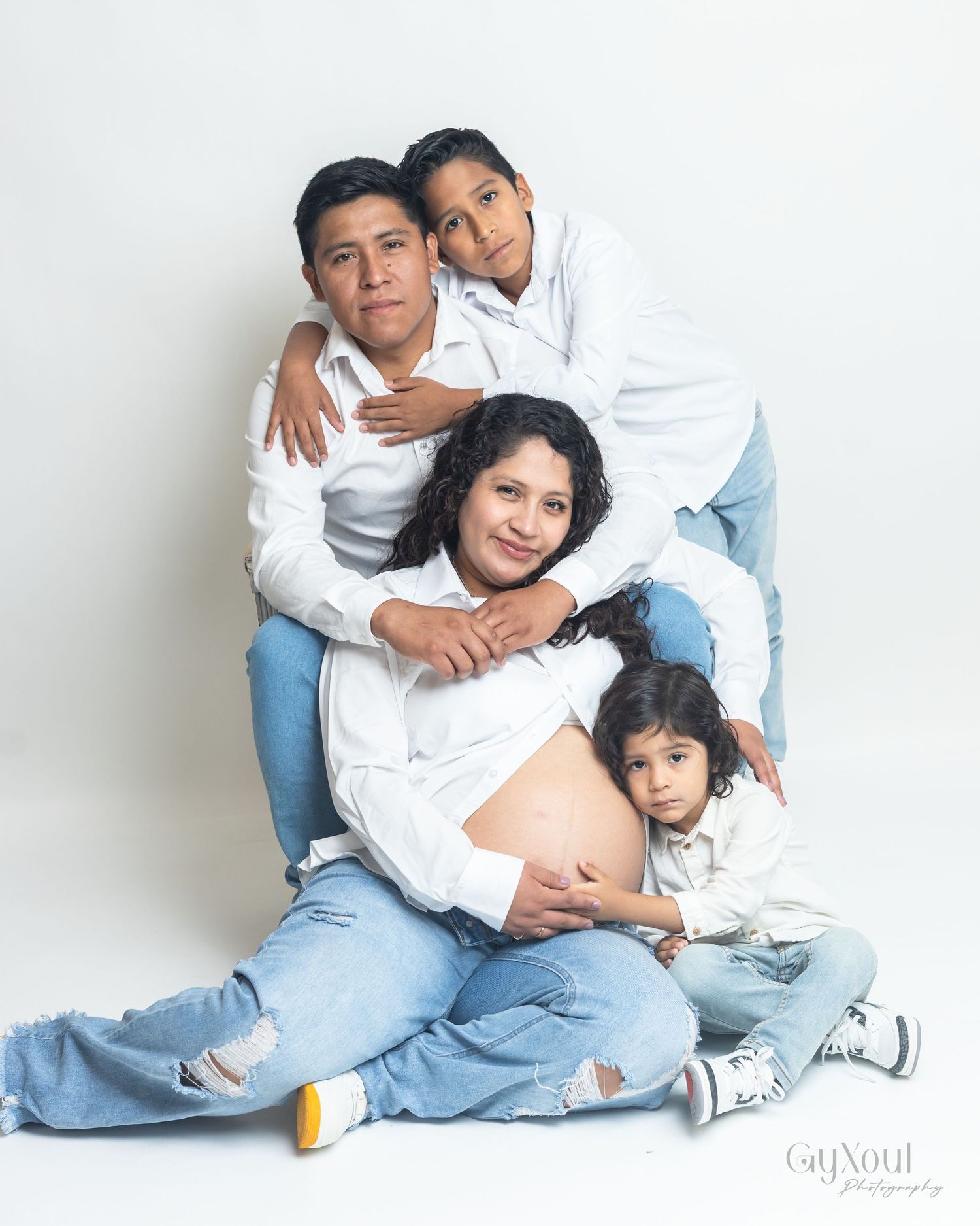 A family wearing matching white shirts and blue jeans poses together against a white background in a studio portrait.