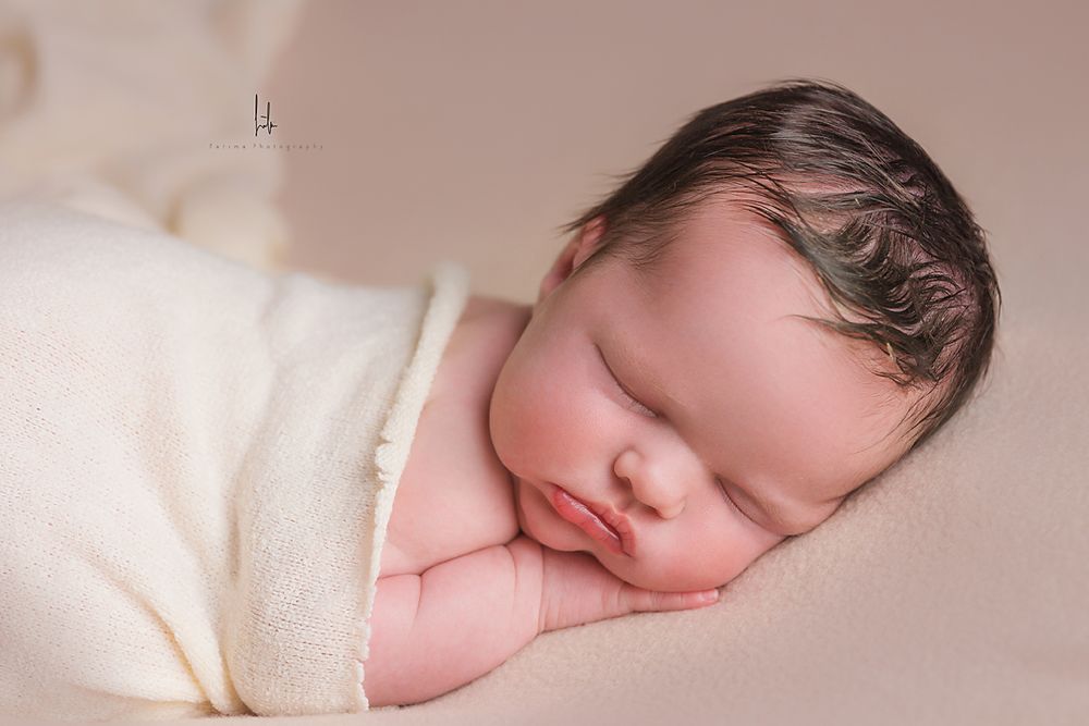 Peaceful sleeping newborn wrapped in soft white blanket against neutral beige background.