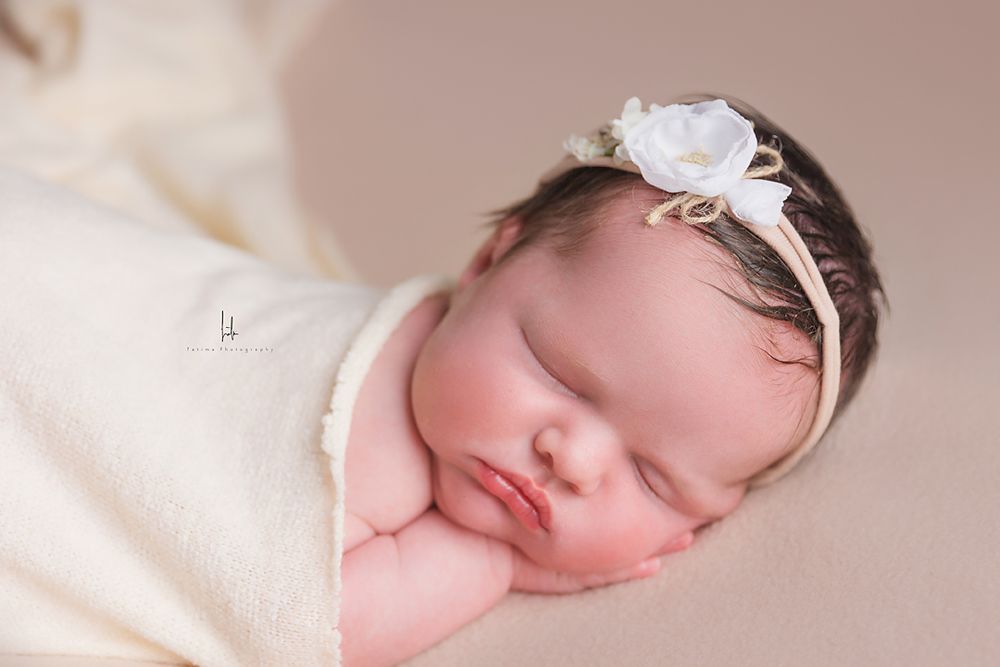 Sleeping newborn wearing delicate white flower headband and swaddled in cream blanket.