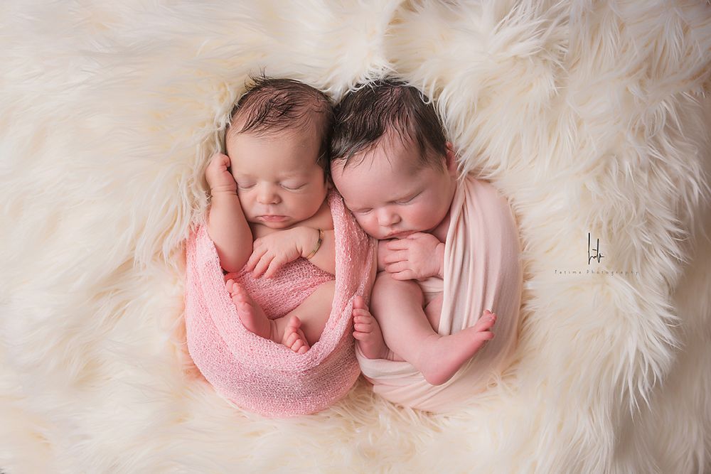 Two sleeping newborn babies wrapped in pink and white swaddles lying together on soft white fur.