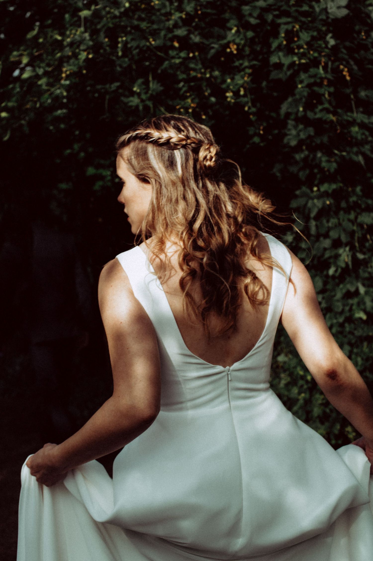 A person in a white sleeveless dress shown from behind in a series of artistic back view poses against dark foliage.