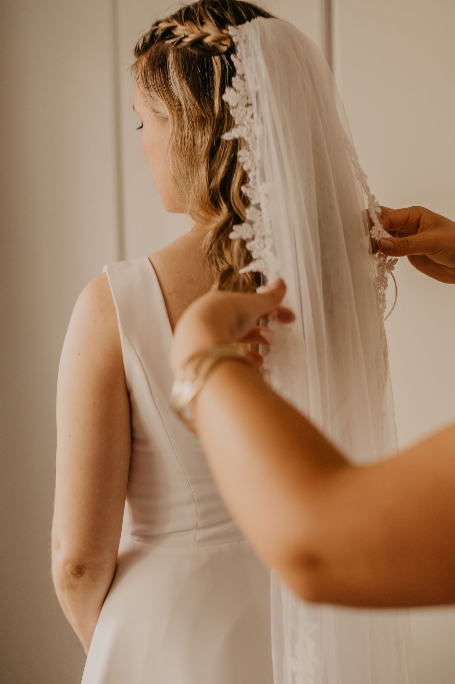 A wedding veil being adjusted on a bride wearing a sleeveless white dress in a warm-toned setting.