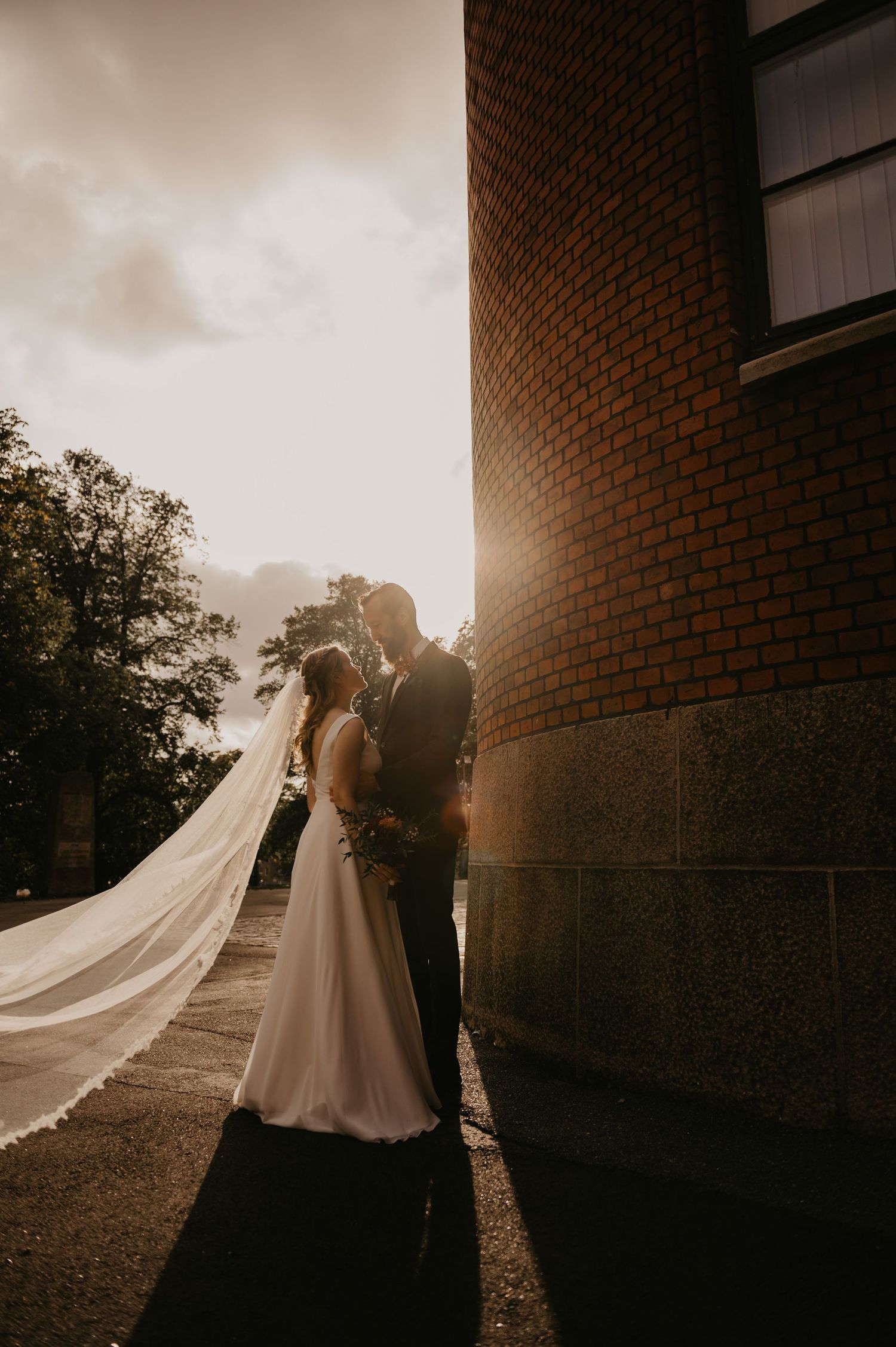 Silhouetted couple shares intimate moment at sunset against brick building as bride's veil flows dramatically in the breeze.