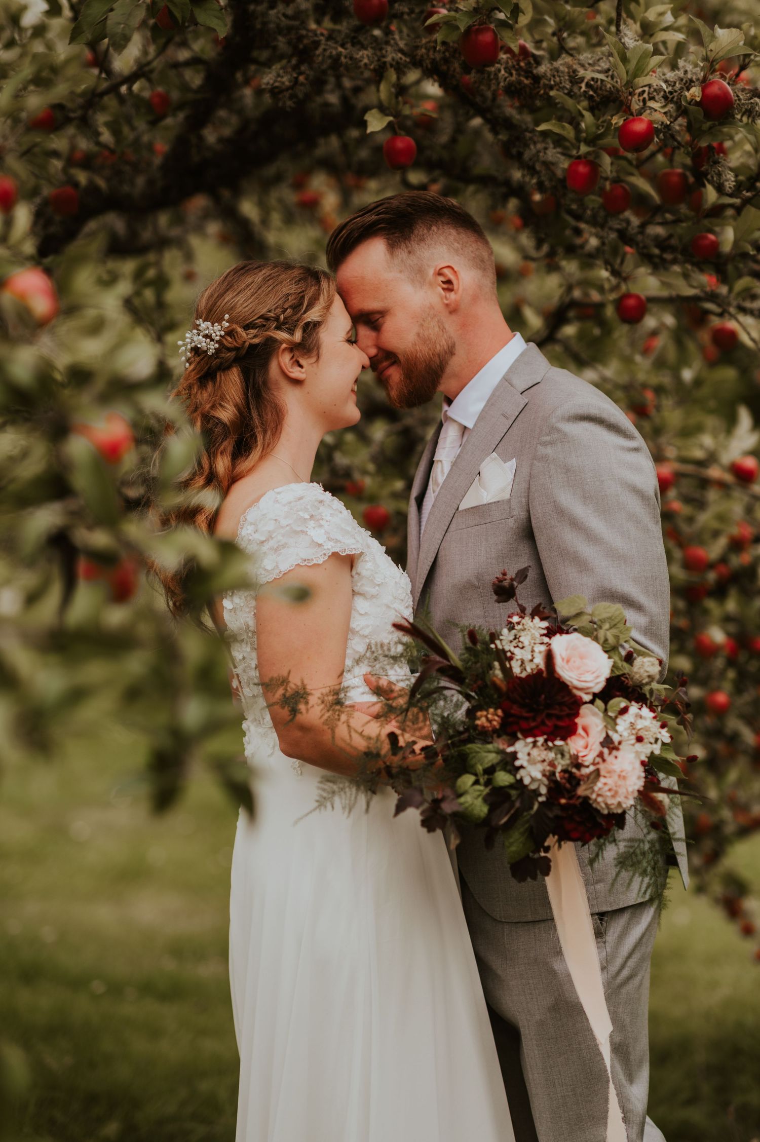 A romantic moment between newlyweds in a dreamy apple orchard with soft natural lighting and pink floral bouquet details.