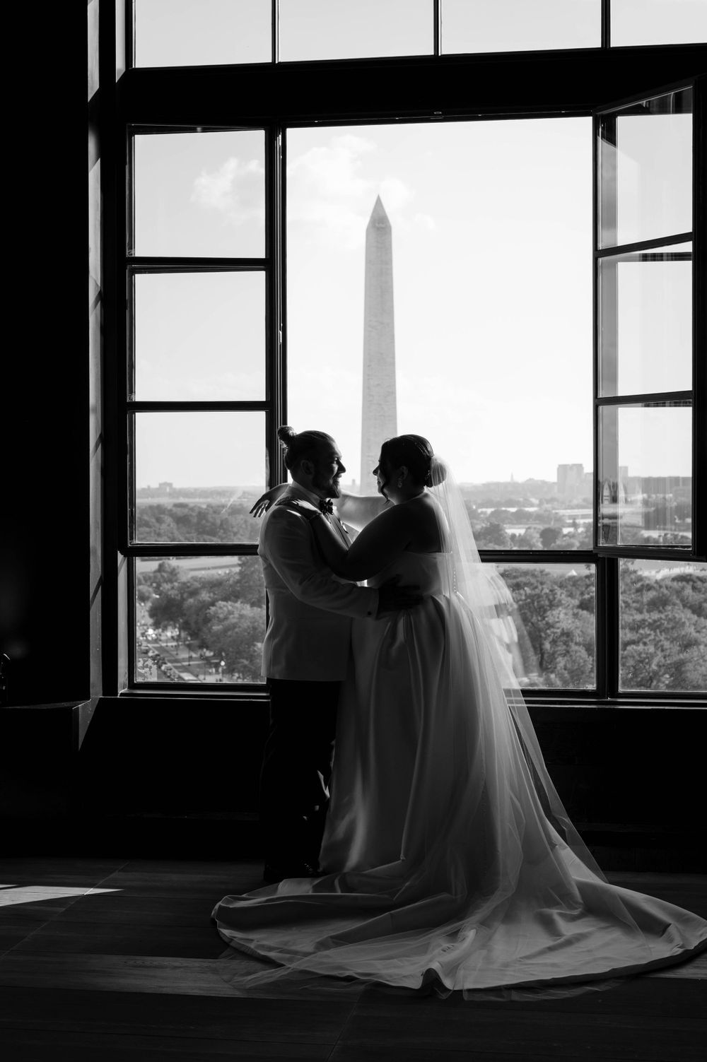 Silhouetted figures in wedding attire gaze through large windows at a scenic cityscape view in dramatic black and white.