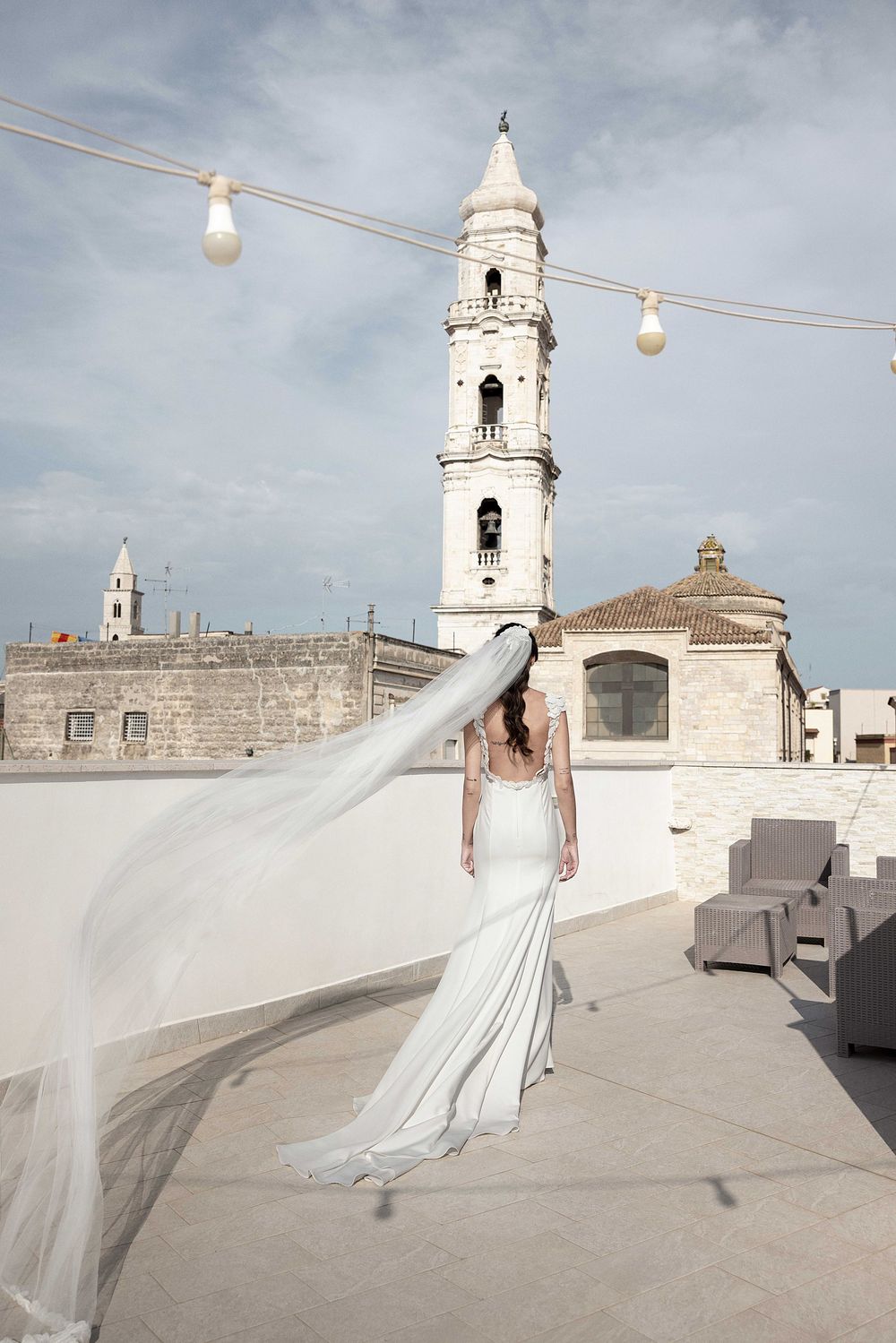 A bride in a flowing white wedding dress with a long veil poses on a rooftop with a church bell tower in the background.