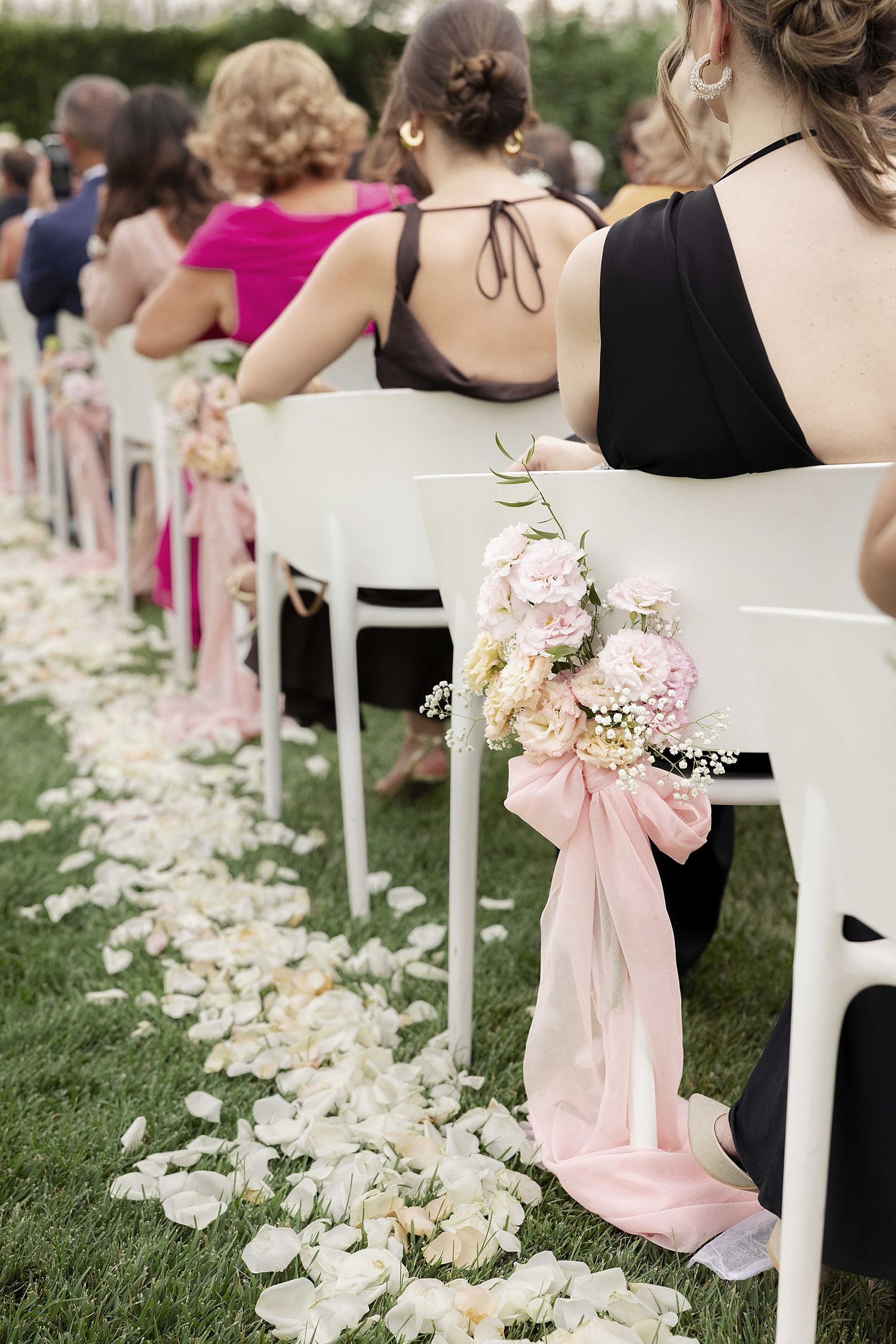 Pink satin bows and scattered rose petals decorate white chairs along an outdoor wedding ceremony aisle.