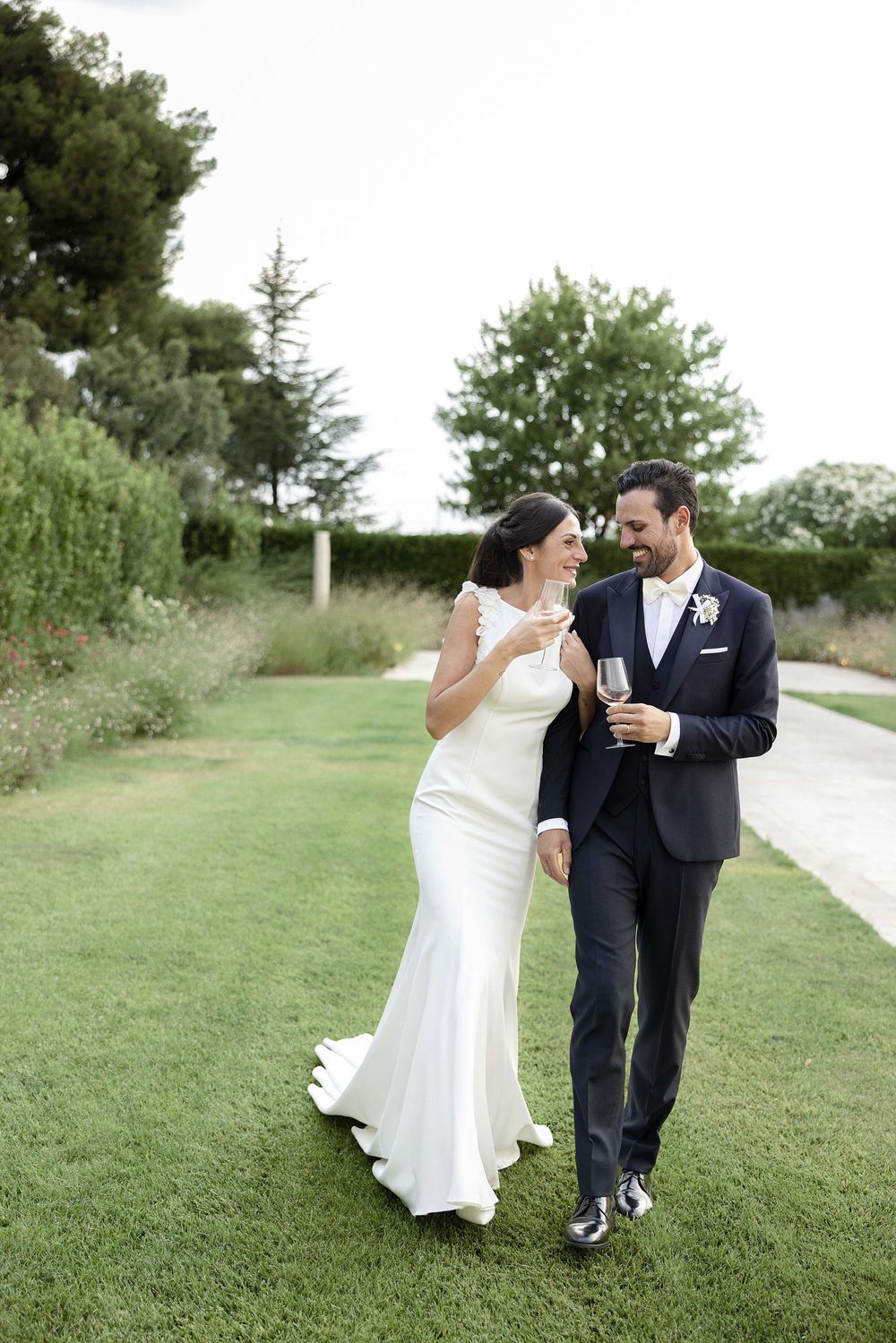A couple in wedding attire walking together and sharing drinks on a garden path surrounded by lush greenery.