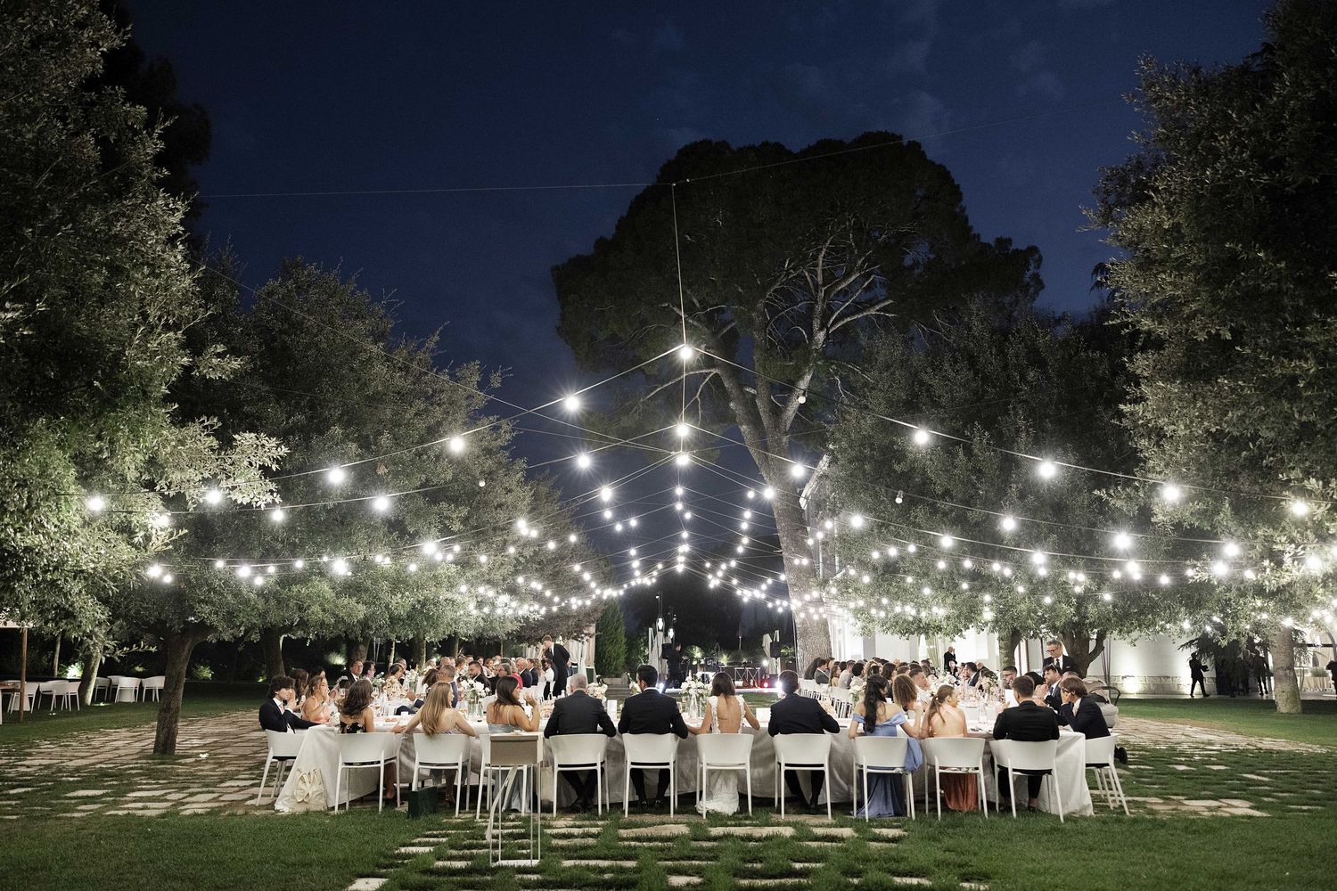 String lights illuminate an outdoor wedding reception at night with tables set under a canopy of trees.