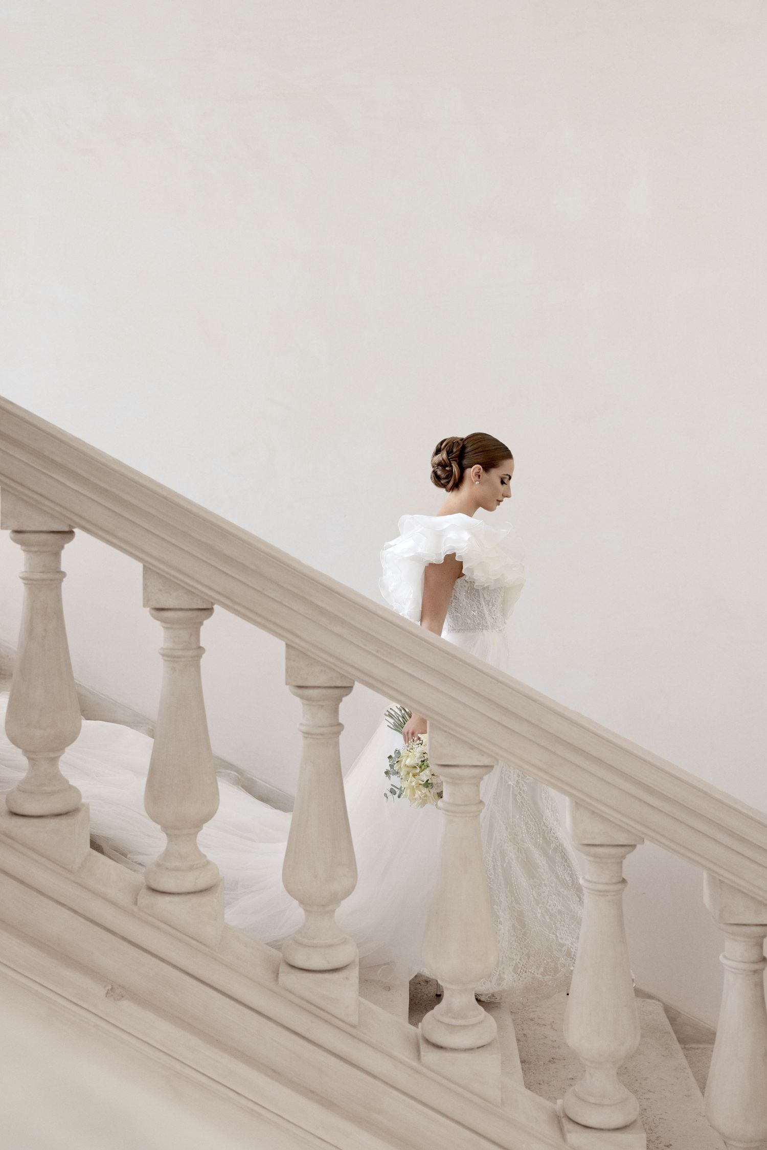 A bride in a white gown gracefully descends a grand staircase with ornate white balusters.
