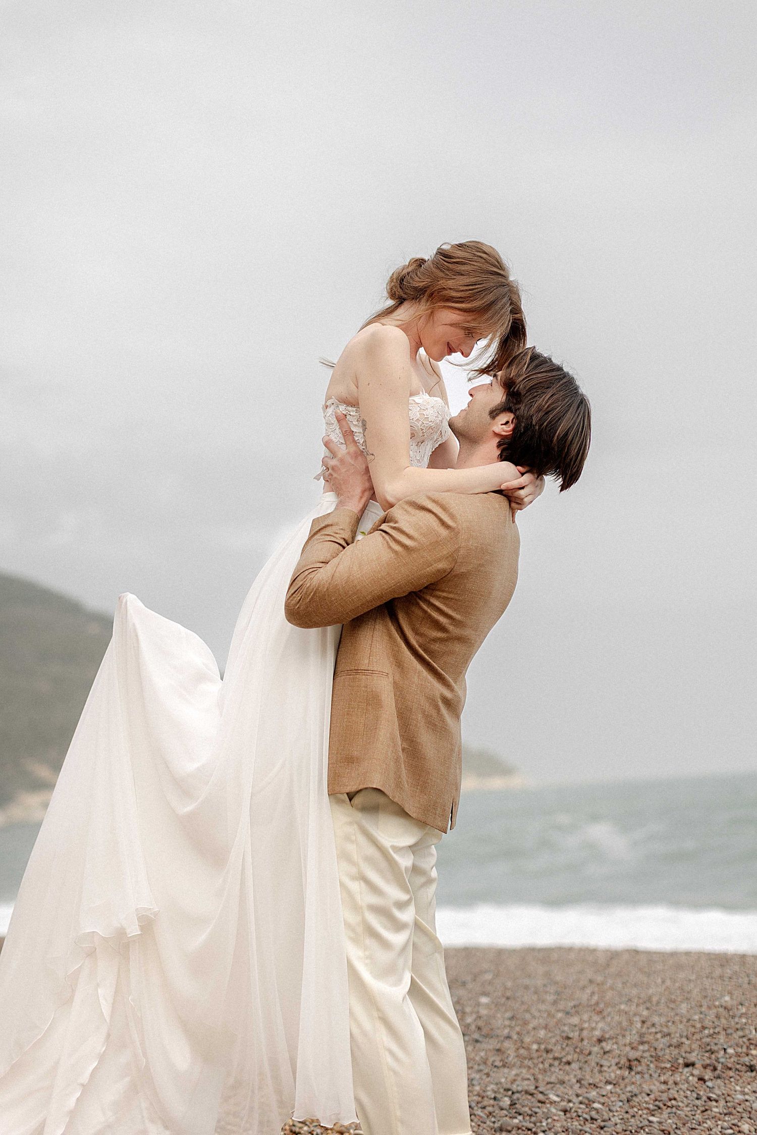A couple shares a romantic embrace on a rocky beach with flowing white fabric and tan attire against a misty backdrop.