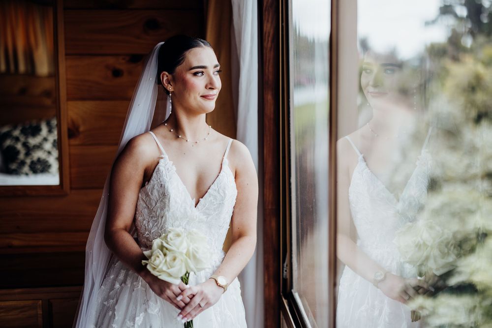 Bride holds white floral bouquet while posing by window in elegant lace wedding gown.