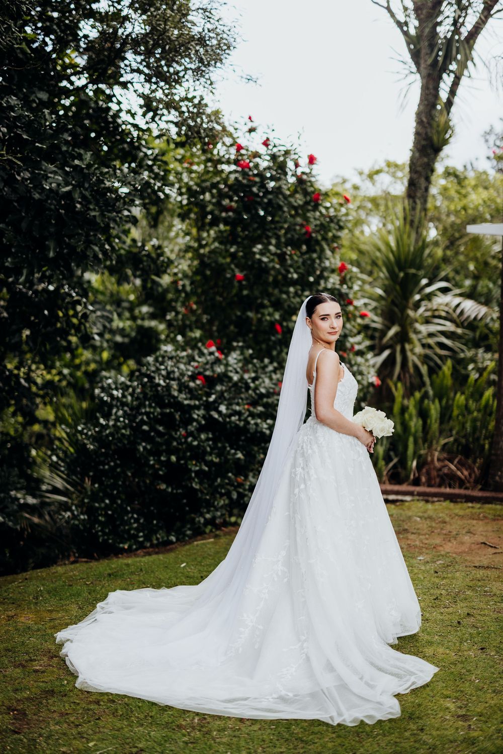 Bride in flowing white gown poses in garden setting with red flowers and palm trees.
