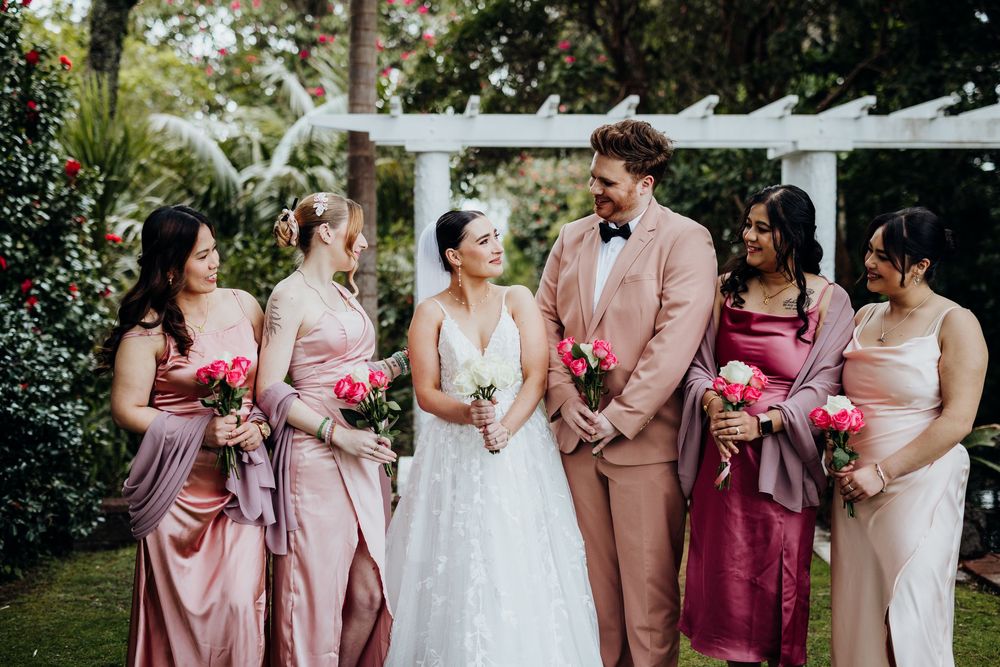 Wedding party poses outdoors wearing pink and burgundy attire with white floral accents.