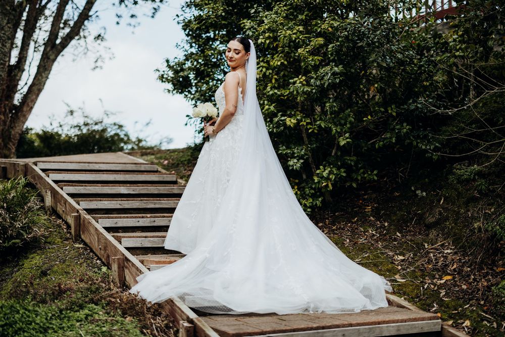 A bride in a flowing white wedding gown poses outdoors on stone steps surrounded by greenery.