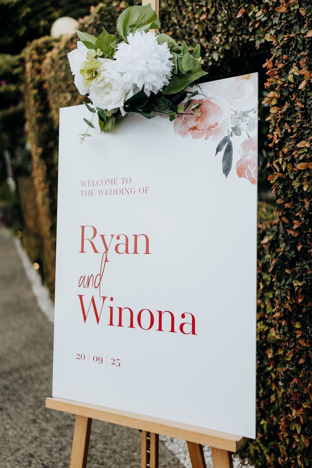 White wedding welcome sign with floral decorations displays names and date on an easel.