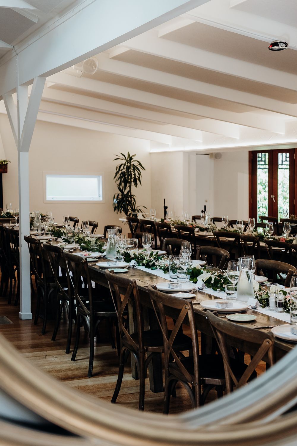 An elegant wedding reception venue with long wooden tables and white decor in a bright indoor space.