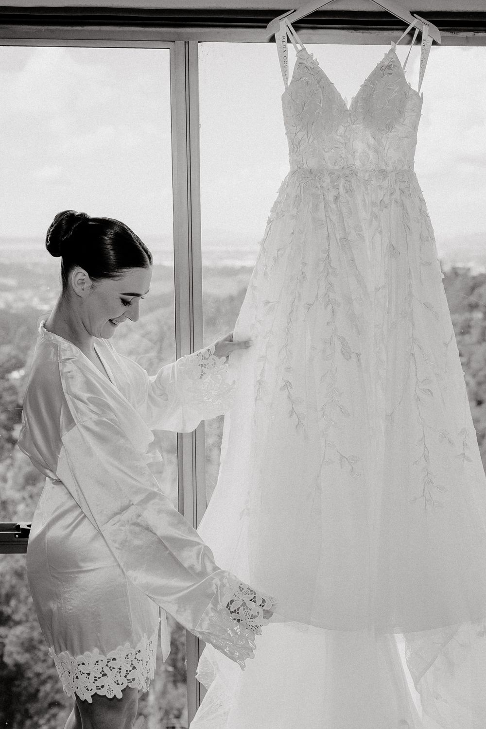 Elegant white lace wedding dress hanging in window light while bride in robe prepares for her special day.