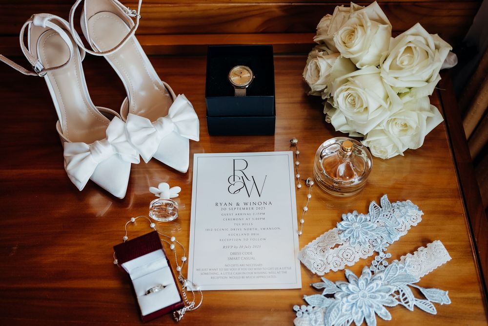 Wedding accessories including white shoes, invitation, rings and roses arranged on wooden surface.